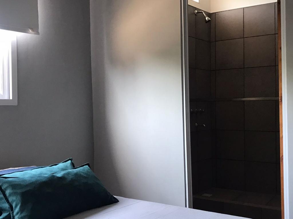 Dormitorio del dúplex con cama y almohadón verde, junto a una ducha con mampara de vidrio